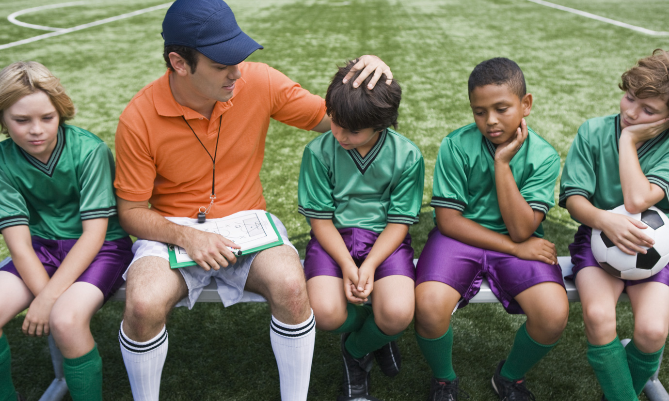 A soccer coach sits on a bench with the disappointed-looking boys on his team and encourages them.