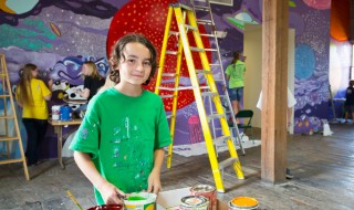 A smiling boy holding paint and looking at the camera as students paint a mural together behind him.