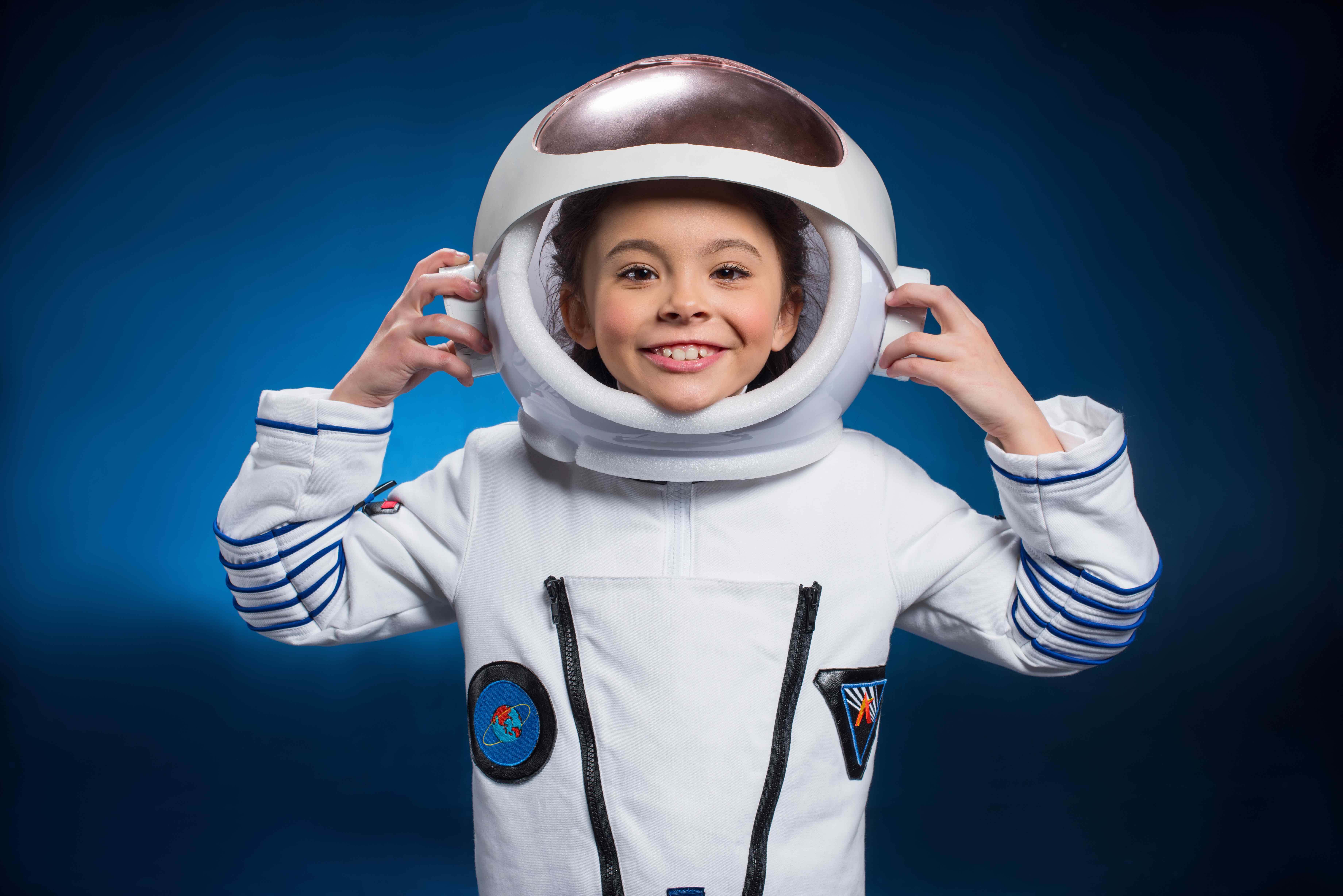 A girl wears a space suit and smiles are she holds up her space helmet.