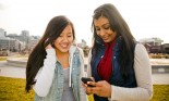 Two young girlfriends (14-17 years old) smiling and using cellular devices together outside in a park, Seattle, Washington