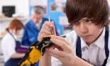 Student (13-15 years old) working on robotic device in vocational class; girl and teacher in background