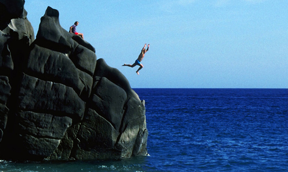 Young man jumping from rocky cliffs into the ocean