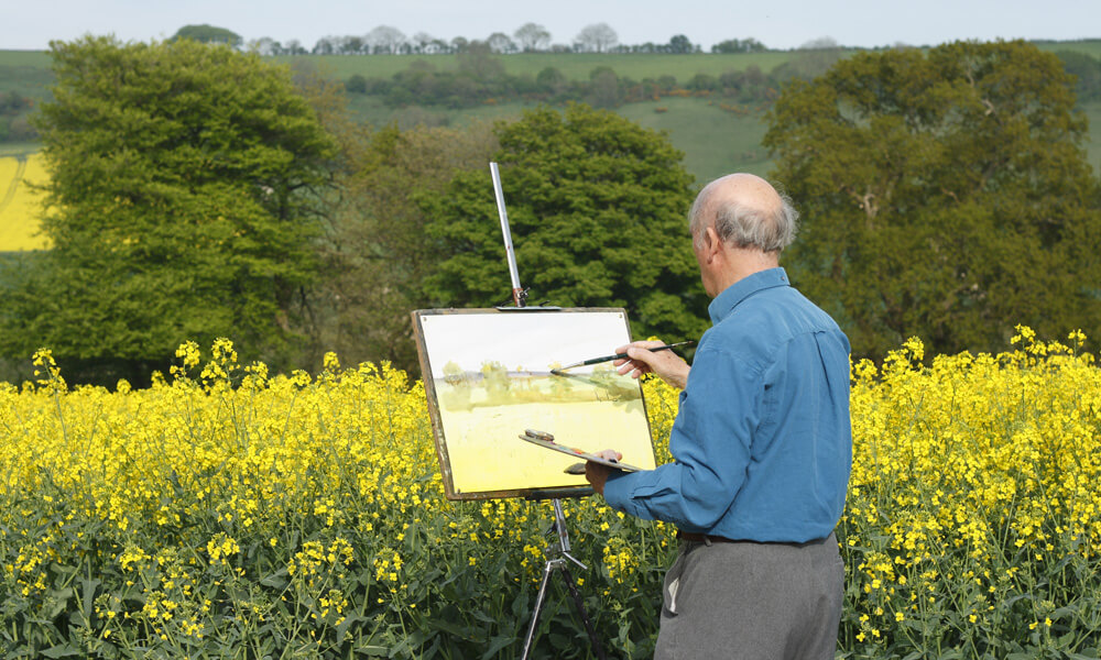 Senior male artist painting in a field full of bright yellow flowers