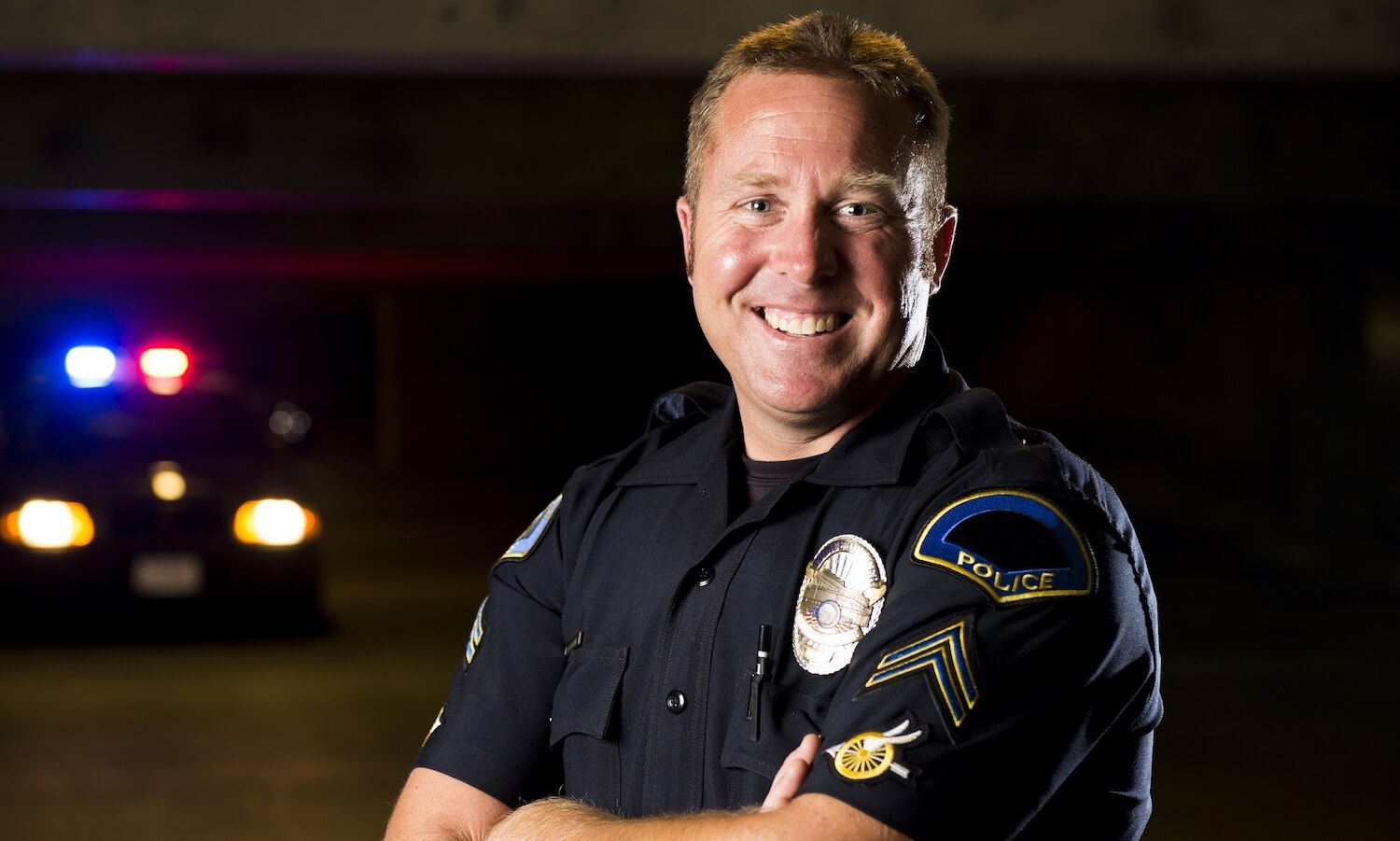 A smiling police officer with his patrol car in the background.
