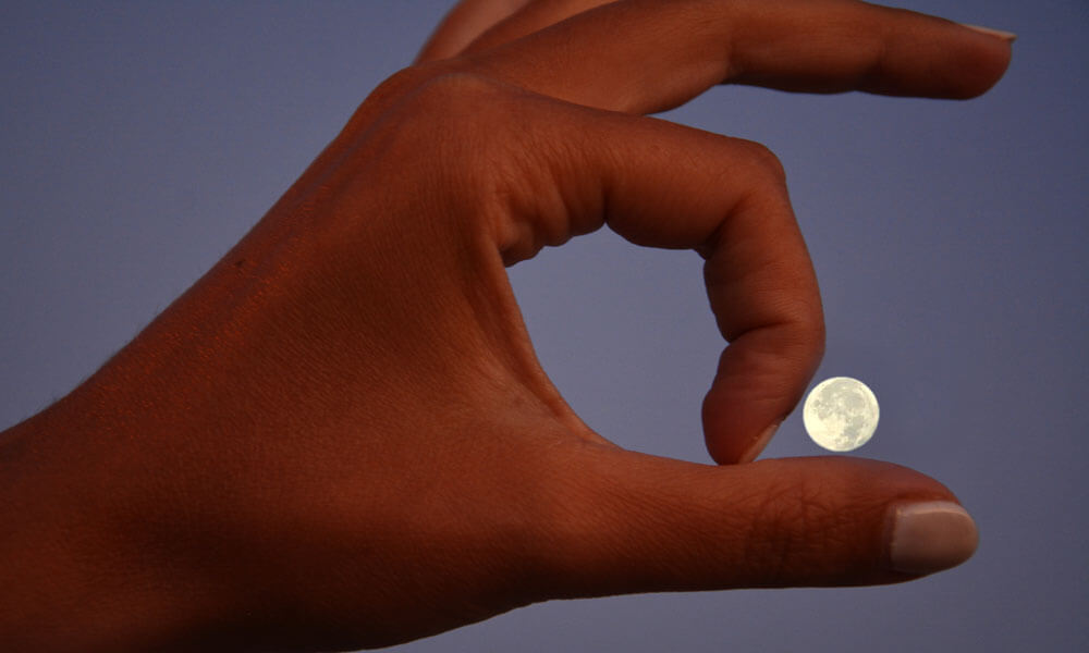 Photo of a hand held up in front of the moon so that it looks like the moon is resting between the thumb and index finger