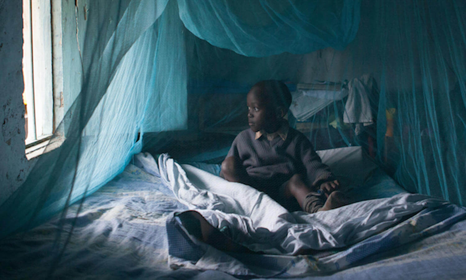 A child sits on a bed underneath a mosquito net near an open window in Kenya, Africa