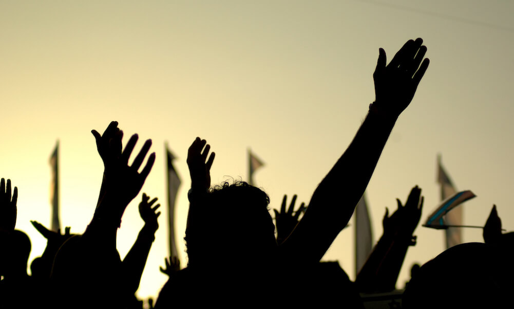 A crowd of people in silhouette raises their hands against the background of a yellow sunset