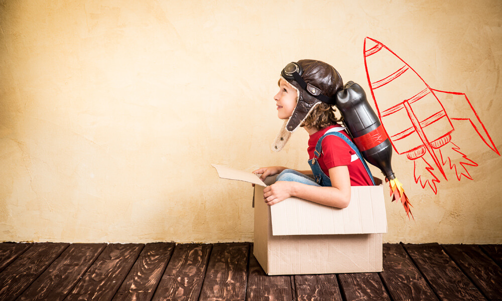 Child sitting in cardboard box wearing jetpack.