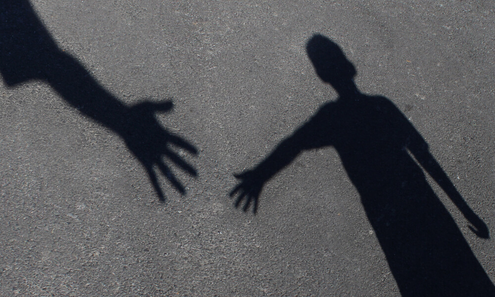 Photo showing the shadow of a hand reaching out to the shadow of a person who is reaching out for help