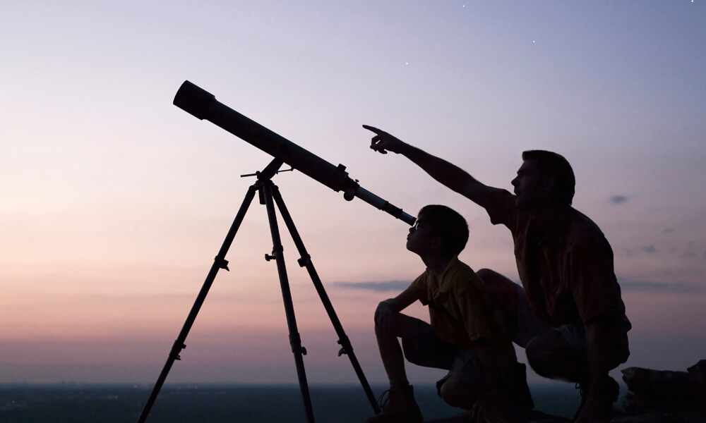 Silhouette of father and son looking through telescope at sunset