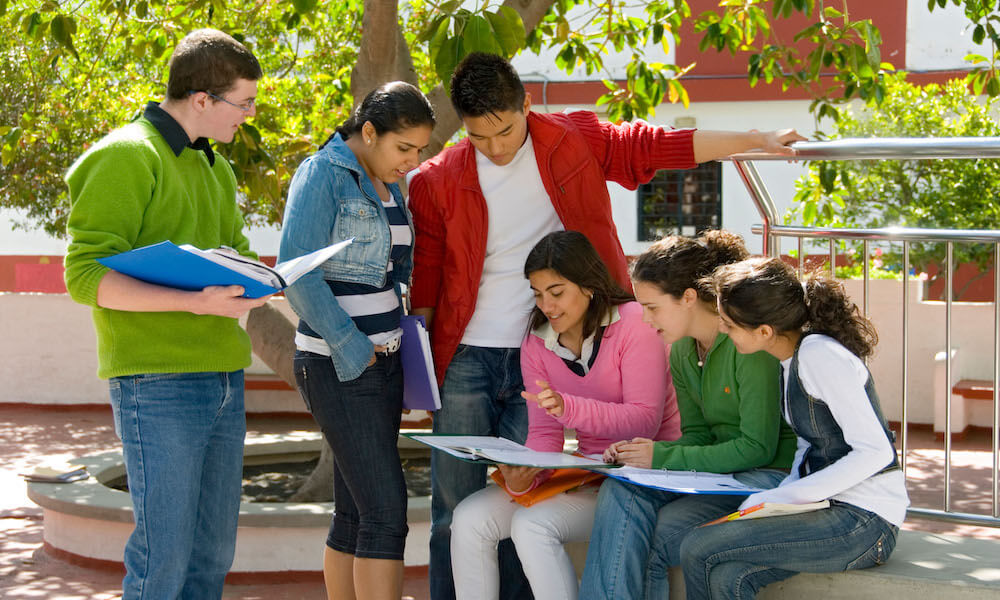 Multiracial group of six senior teenage students studying outdoors and comparing notes together