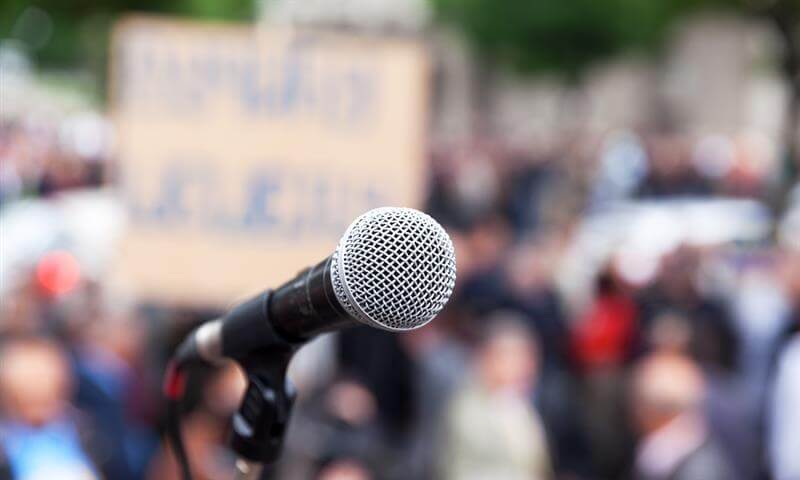 Microphone with blurred audience of protestors and a sign in the background.