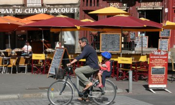 A man and child ride a bicycle in front of cafes on the the Rue Champ Jacquet in Rennes, France.