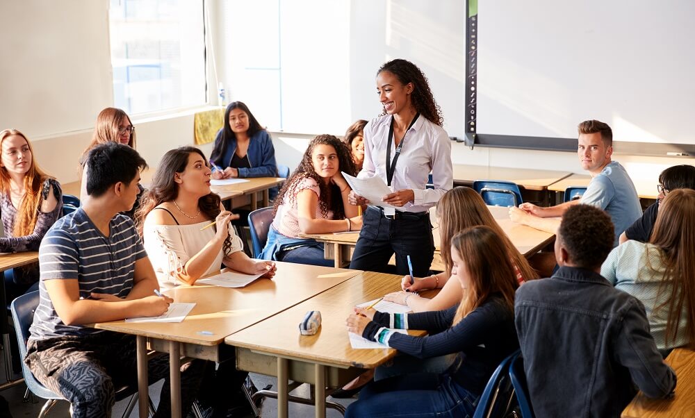 Female High School Teacher Standing By Student Table Teaching Lesson