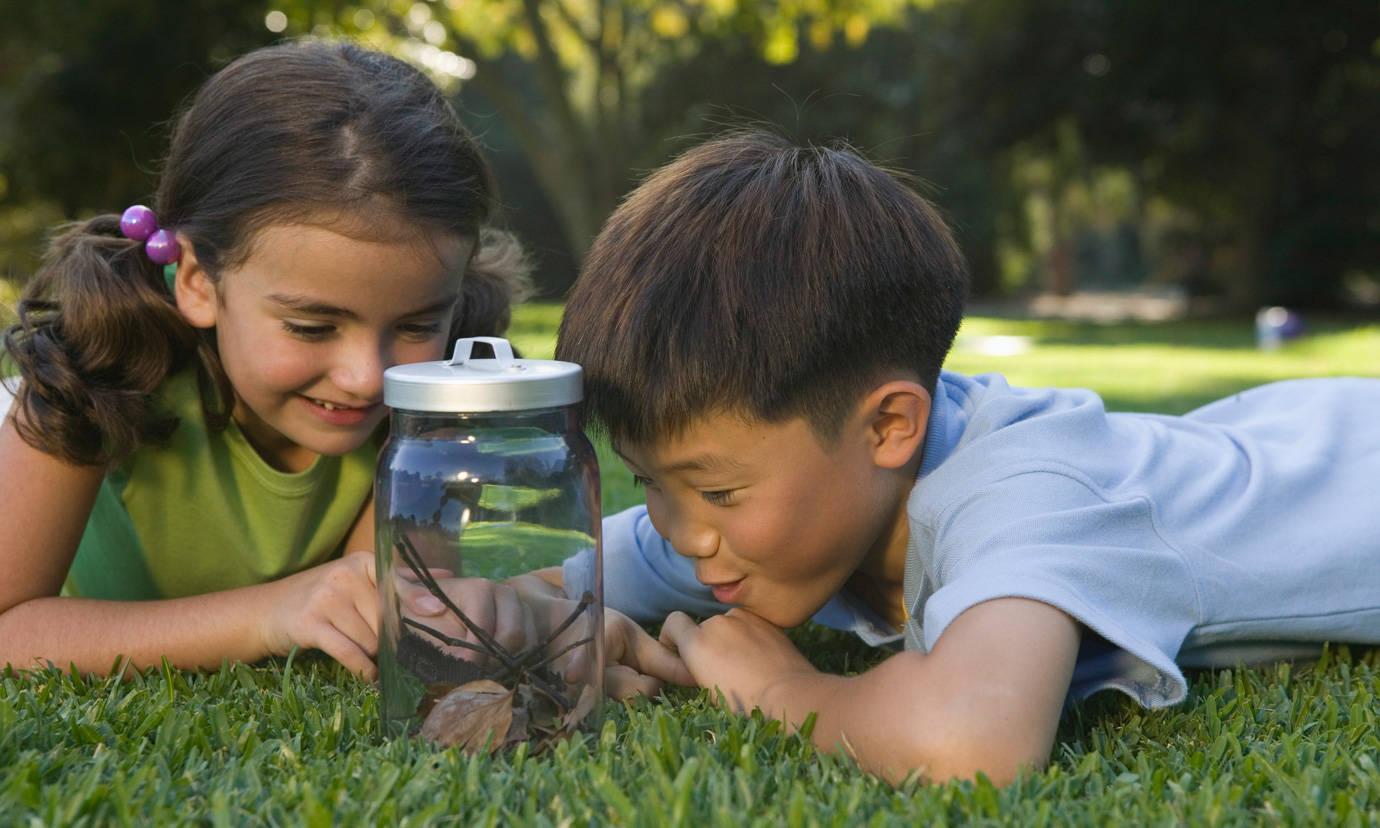 A boy and girl lying on grass in a park, looking at a glass jar holding some leaves and twigs.