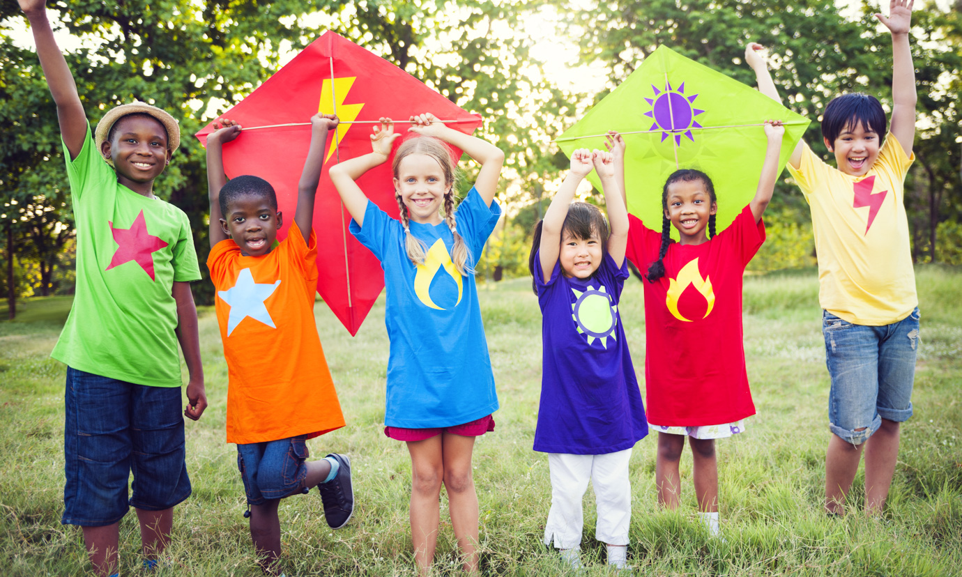 A group of kids in a grassy field, holding two colorful kites over their heads.