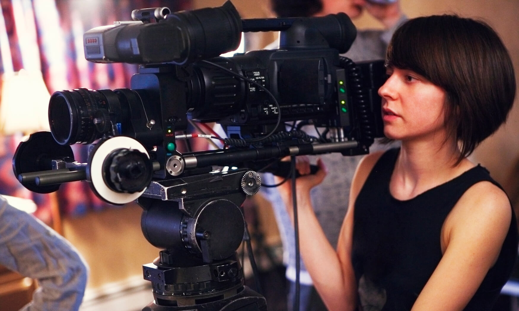 A young woman operating camera on a video shoot. Camera has a 35mm adaptor on it.