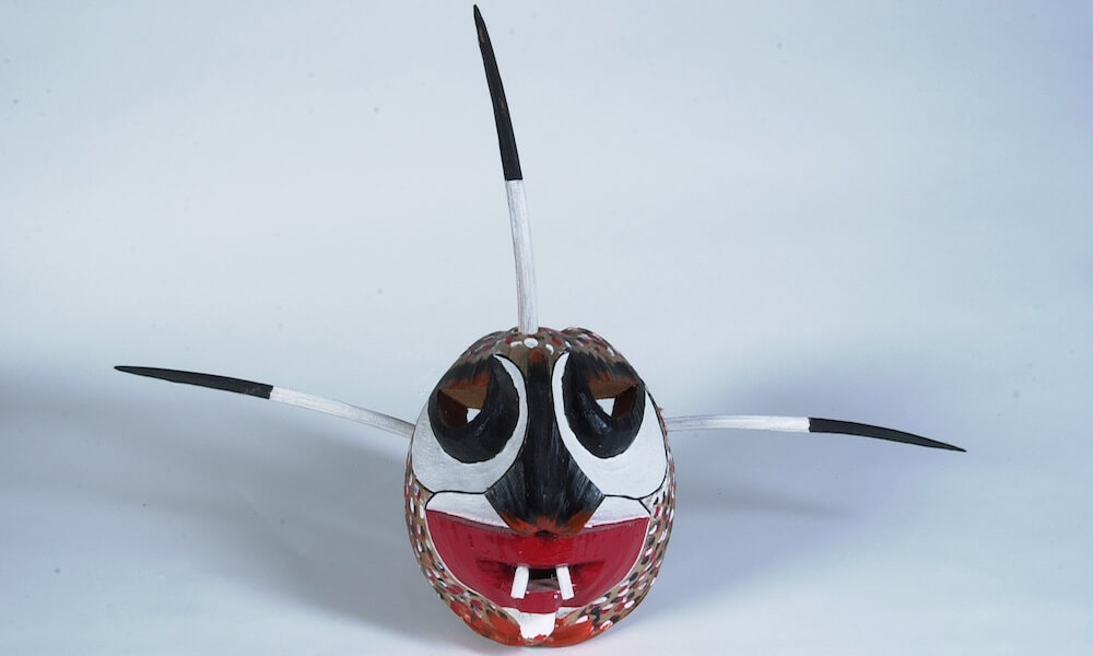 Una máscara vejigante, hecha de coco. Tiene una cara con ojos negros y una boca roja con dos dientes. Tres cuernos salen de la cabeza.