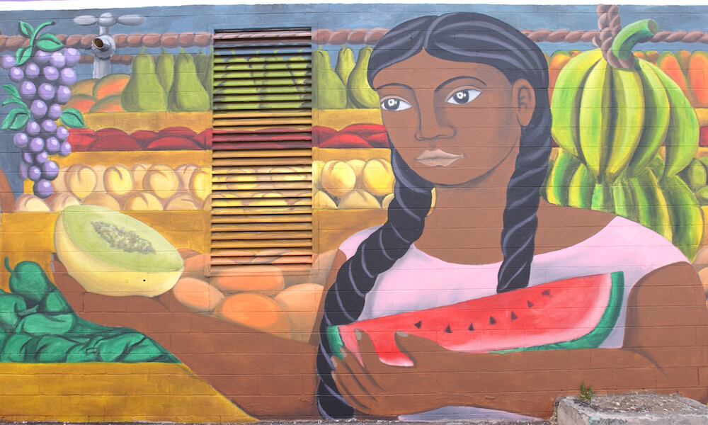 Una vendedora de frutas morena lleva un sandía en un mural pintado en una pared