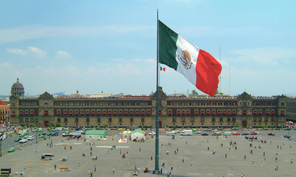 La bandera mexicana ondea en medio de la Plaza de la Constitución, en la Ciudad de México. Al fondo se ve el Palacio Nacional.