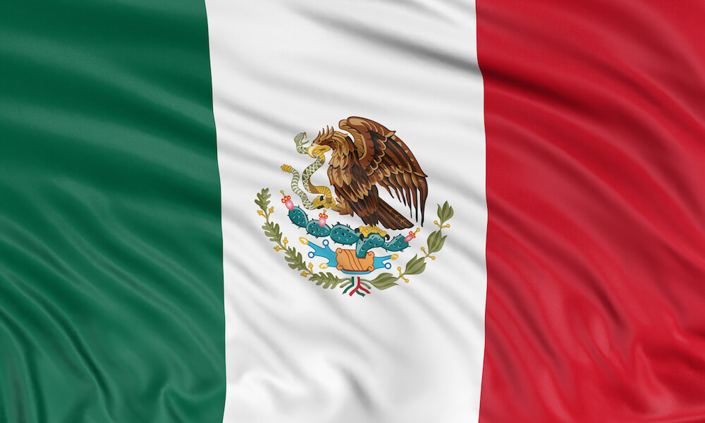 La bandera mexicana tiene tres franjas verticales de color verde, blanco y rojo. En el centro, en medio de la franja verde, se ve el escudo nacional que tiene un águila que come una serpiente mientras se posa en un nopal.