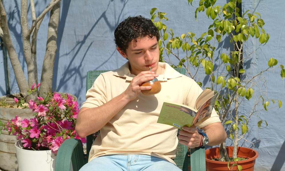 A young man drinks mate and reads a book while sitting on an outdoor patio.