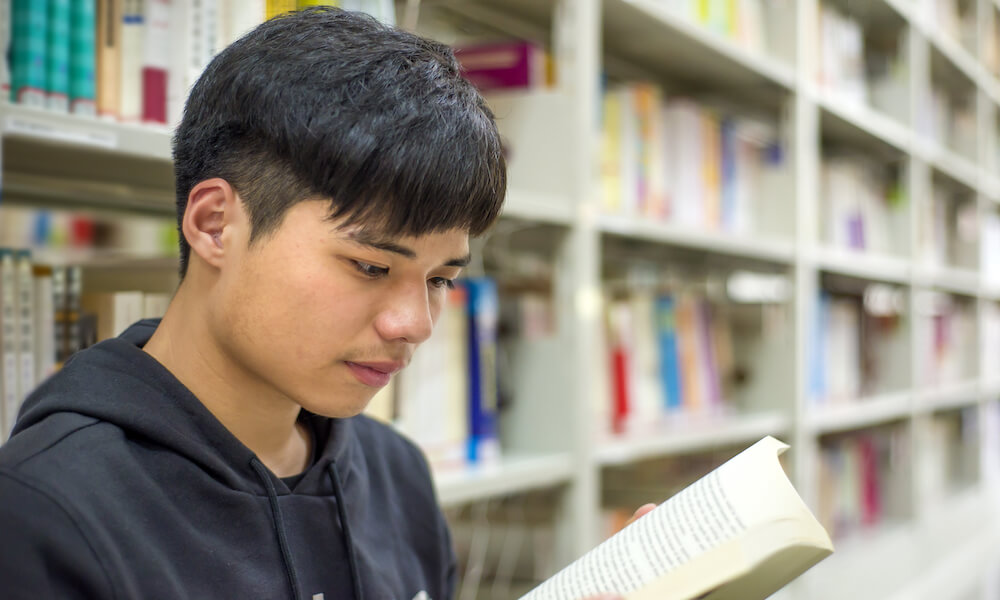 A teenage boy reads a book in the stacks of a library.