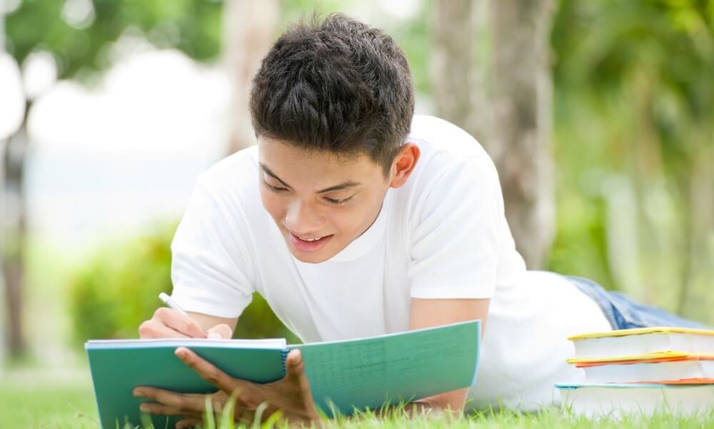 Male college student lying on the grass and writing in notebook